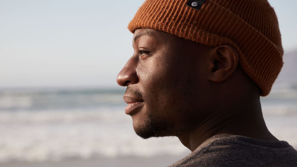 A person wearing a rust-colored knit beanie looks out toward the ocean with a neutral expression, perhaps reflecting after counseling. The background is a blurred view of the sea and sky.